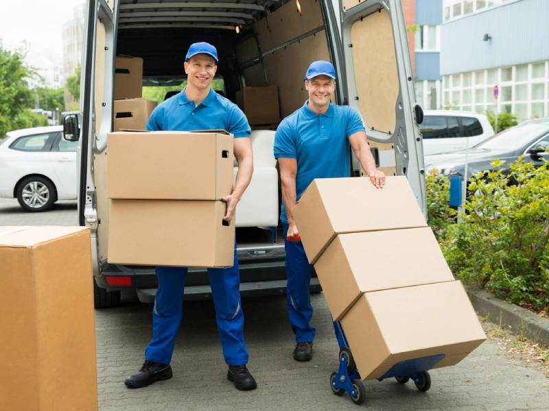 Two men wearing blue, smiling while unloading boxes from a van
